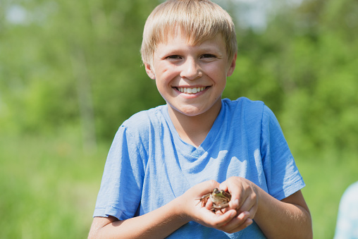 An adorable elementary school boy of Caucasian ethnicity is holding a toad in his hand. He is outdoors on a warm summer day.