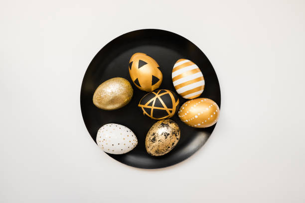 istock Stylish Easter golden decorated eggs on black plate isolated on white background. Trendy flat lay easter. Happy Easter card with copy space for text. Minimal easter concept 1301302132