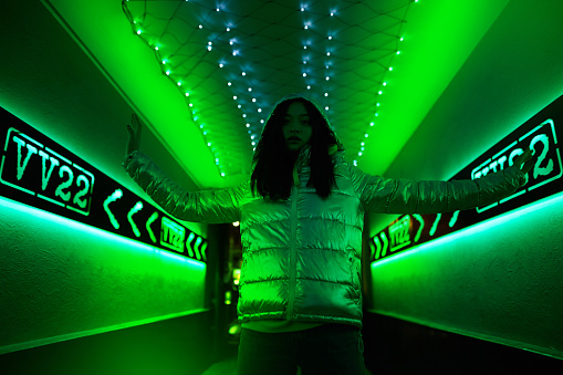 Nightlife and party concept. Low angle view of confident and trendy young adult girl standing in club with bright green neon light
