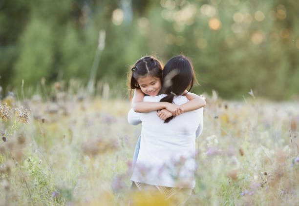 A little girl around 7 years old is hugging her mother as they are both standing outdoors in a meadow.