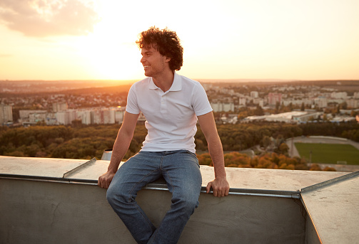 Positive young man with curly hair smiling and looking away while sitting on roof and enjoying freedom against city and sundown sky