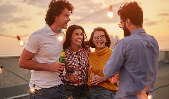 Group of happy young people in casual clothes chatting and laughing while drinking beer and having fun during party, on rooftop of urban building at summer sunset