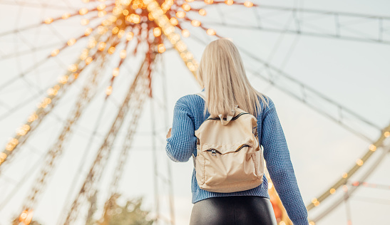 From below back view of unrecognizable blond female in casual clothes with backpack looking away while standing alone, against illuminated Ferris wheel and blue sky on fairground