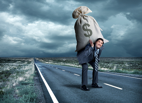A businessman struggles as he carries a large money bag on his back on a long straight rural road under ominous skies.