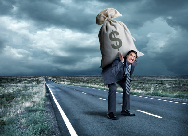 Man Struggles To Carry Money Bag On His Back A businessman struggles as he carries a large money bag on his back on a long straight rural road under ominous skies. greedy corporation stock pictures, royalty-free photos & images
