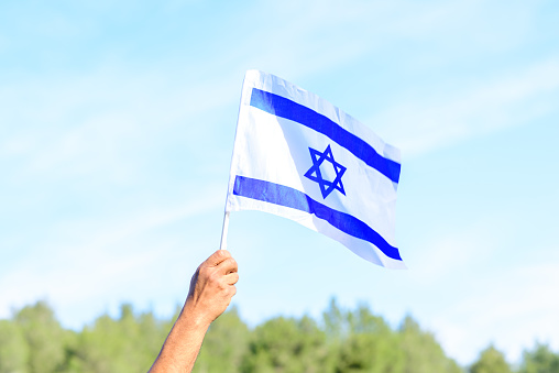 Israeli flag, man hand, blue sky, nature background.Memorial day-Yom Hazikaron, Patriotic holiday Independence day Israel - Yom Ha'atzmaut concept.