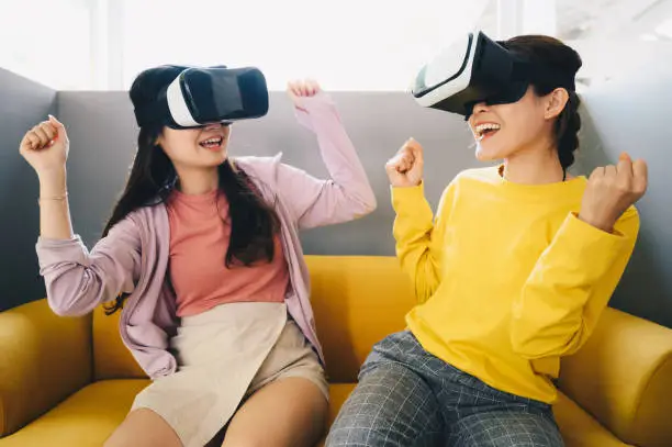 Photo of Two young Asian women wearing virtual reality glasses and playing video games together.