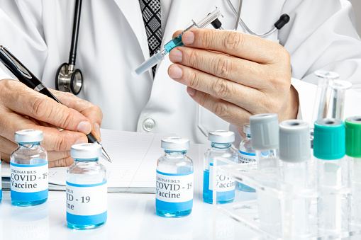 A doctor holding a glass syringe writing the test result on a notebook with more vials labelled covid-19 vaccine and some test tubes in a rack on a white table. Covid-19 vaccine testing concept.