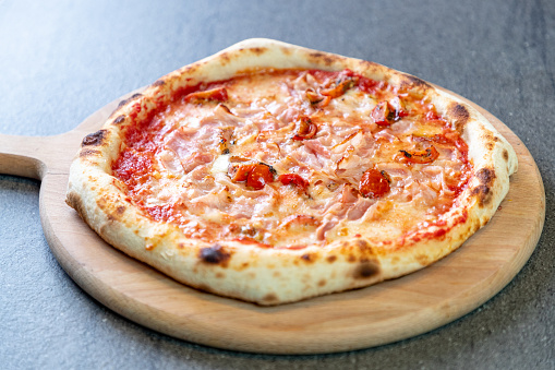 Baked pizza with tomato sauce base, garnished with bacon, mozzarella cheese and cherry tomato chops on round wooden board