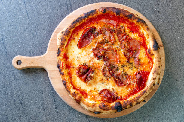 Directly above pizza garnished with mozzarella cheese and red prosciutto slices on tomato sauce, seasoned with basil, served on round wooden board