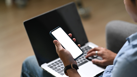 Close up view of casual man working on laptop and using mobile phone while sitting on couch at home.