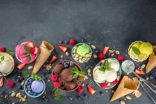 Various colorful ice cream balls in different bowls, with ice cream waffles cones and flavor ingredients - pistachio nuts, berries, lemon, chocolates, vanilla beans, mint. Dark background copy space