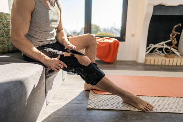 Young man putting on knee bandage fixator before exercising Young man sitting on sofa and putting on knee bandage fixator before exercising knee brace stock pictures, royalty-free photos & images