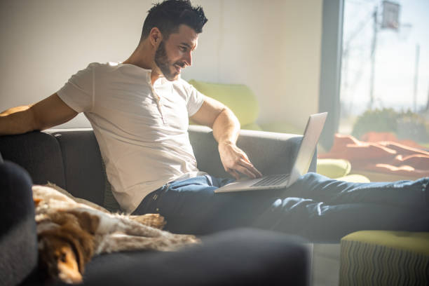 istock Young bearded man in tshirt sitting on sofa and using laptop 1301356616