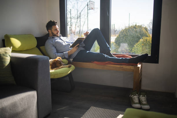 istock Young man lying on bench next to window and reading book while his dog lying next to him 1301358014