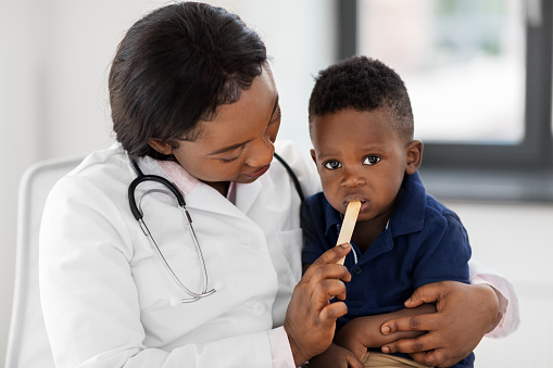 medicine, healthcare, pediatry and people concept - african american female doctor or pediatrician with stick examining baby boy patient's mouth at clinic
