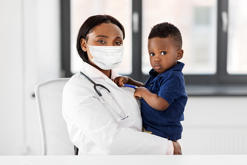 medicine, healthcare, pediatry and people concept - african american female doctor or pediatrician wearing protective mask holding baby boy patient on medical exam at clinic