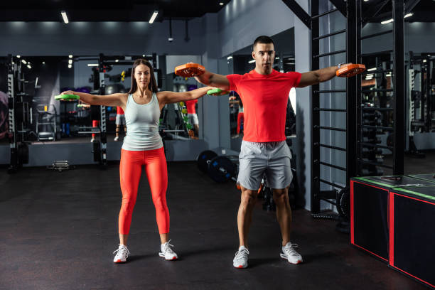 A muscular man and a beautiful young woman doing some arm and shoulder exercises together in the indoor gym at night time. Couple goals, fitness goals A muscular man and a beautiful young woman doing some arm and shoulder exercises together in the indoor gym at night time. Couple goals, fitness goals running shoes on floor stock pictures, royalty-free photos & images