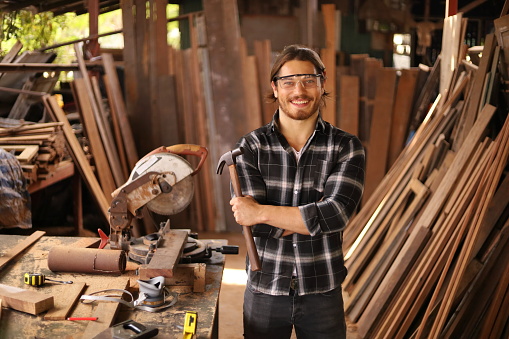 Portrait of smiling young Caucasian carpenter man is working in his own craftsman garage style workshop for hobby with copy space