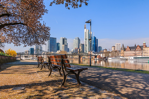 Skyline with skyscrapers of Frankfurt. Financial district with commercial buildings on the day with sunshine. Main riverbank in spring with tree and benches and ships on the pier