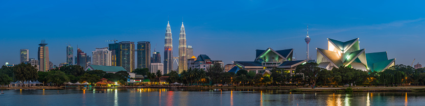 Panoramic view across the landmarks of Kuala Lumpur, from the iconic twin skyscrapers of the Petronas Towers spotlit against the deep blue dusk sky to the soaring spire of Menara Kuala Lumpur and Istana Budaya reflecting in the waters of Titiwangsa, Malaysia.