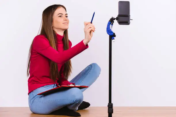 Photo of Study online during quarantine. Young brunette girl during a video conference sitting on the floor with a folder on a white background.