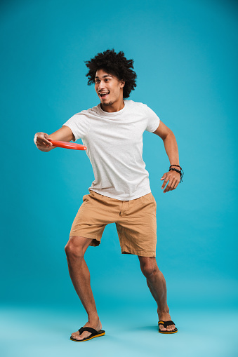 Full length portrait of a happy young african man playing with frisbee isolated over blue background