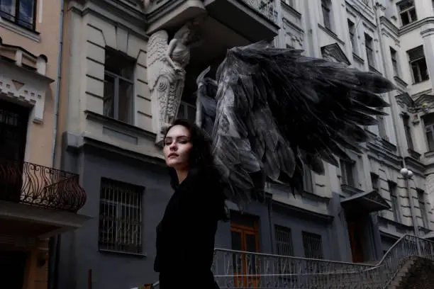 Urban style conceptual portrait of beautiful young woman with dark wings as angel walking in the city streets