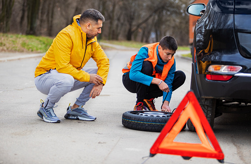 Father showing his son how to change tire.They standing next to the car and changing the punctured tire together.