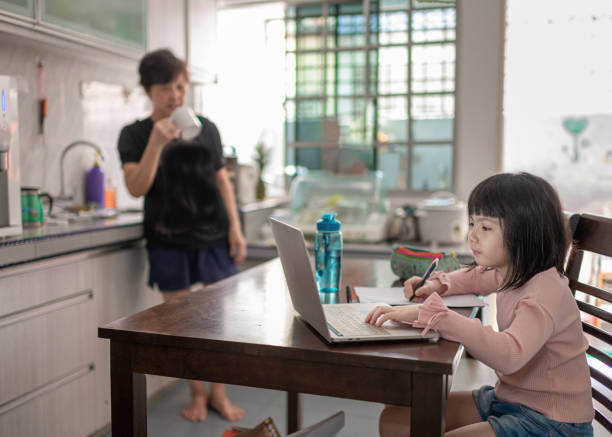 asian chinese young girl attending online class at the kitchen dining table while her grandmother preparing food in kitchen - grandparent using computer laptop dining table imagens e fotografias de stock