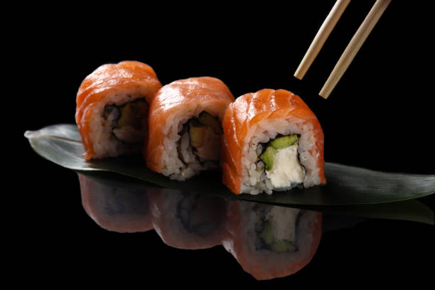 istock Eating sushi rolls with chopsticks. Set on a black background with reflection. Sushi roll with rice, cream chees, salmon, avocado. 1301393463