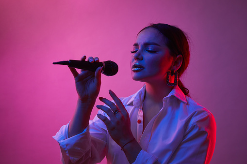 Female singer in neon light. Girl in white shirt with microphone. Concept of art, music.