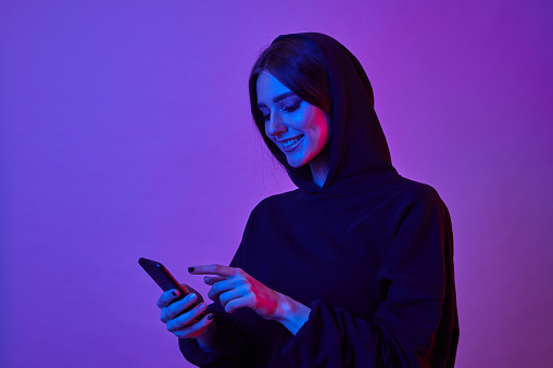 Happy young woman wearing hoodie and using smartphone in neon lights. Girl surfing internet on phone in pink neon light.
