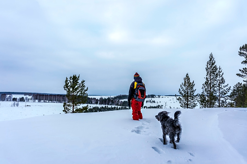 Young man with backpack in winter clothes standing in snow on side of cliff and looking at beautiful winter view and fluffy dog near. Walking and hiking in winter with dog, active lifestyle, landscape