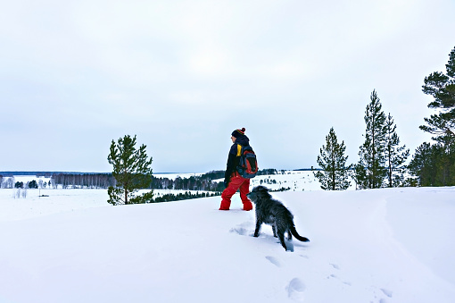 Young man with backpack in winter clothes standing in snow on side of cliff and looking at beautiful winter view and fluffy dog near. Walking and hiking in winter with dog, active lifestyle, landscape