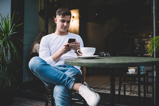 Concentrated young guy sitting with crossed legs and messaging on social media on cellphone while relaxing in cafe with coffee