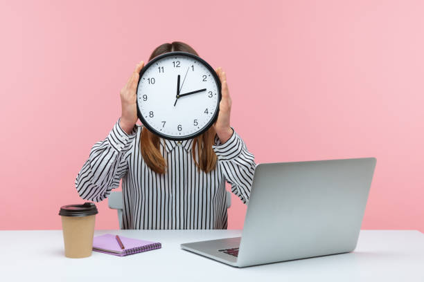 istock Time to work! Woman office worker sitting at workplace hiding face behind big wall clock, time management, schedule and business meeting appointment 1301400356