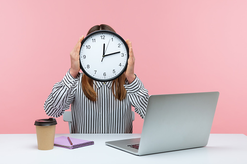 Time to work! Woman office worker sitting at workplace hiding face behind big wall clock, time management, schedule and business meeting appointment. Indoor studio shot isolated on pink background