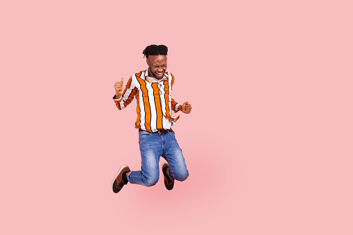 Full length happy positive african man with dreadlocks in stylish bright shirt highly jumping on trampoline, having fun, enjoying like in childhood. Indoor studio shot isolated on pink background