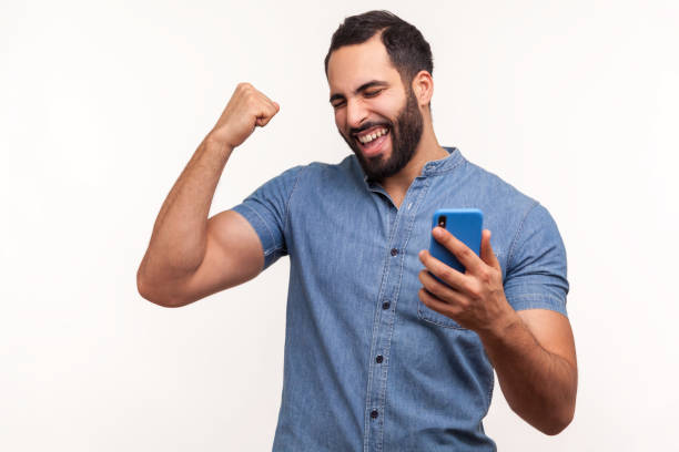 felice uomo soddisfatto con barba in camicia blu che tiene lo smartphone e sorride facendo sì gesto, celebrando lotteria online o vittoria giveaway - latin american and hispanic ethnicity isolated on white happiness cheerful foto e immagini stock