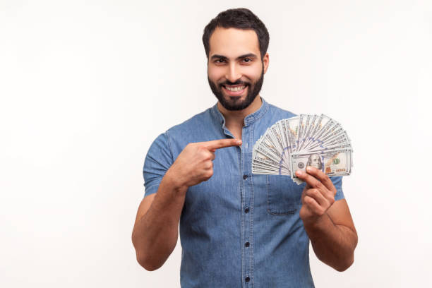 homme d’affaires barbu heureux dans la chemise bleue pointant le doigt à l’argent comptant de dollar dans sa main, regardant l’appareil-photo avec le sourire toothy, bon salaire, paiements - beak buying currency exchanging photos et images de collection