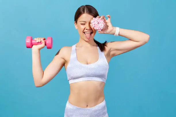 Funny positive sports woman in tights and top having fun, closing eye with donut and showing tongue, holding dumbbell in hand, burning extra calories. Indoor studio shot isolated on blue background