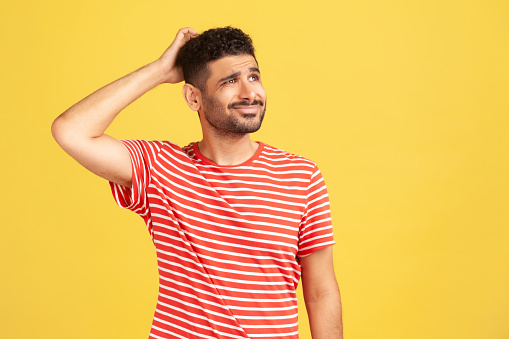 Confused uncertain man with beard in red striped t-shirt scratching his head, choosing, trying to make right decision, dilemma. Indoor studio shot isolated on yellow background