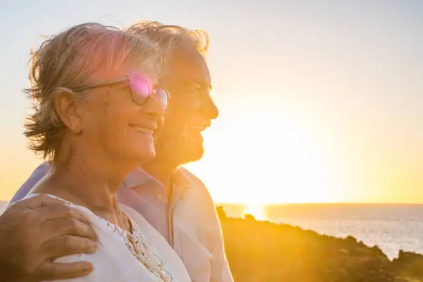 Photo of couple of two cute seniors together enjoying summer and having fun at the beach looking at the sea or ocean with sunset - mature people having a good lifestyle
