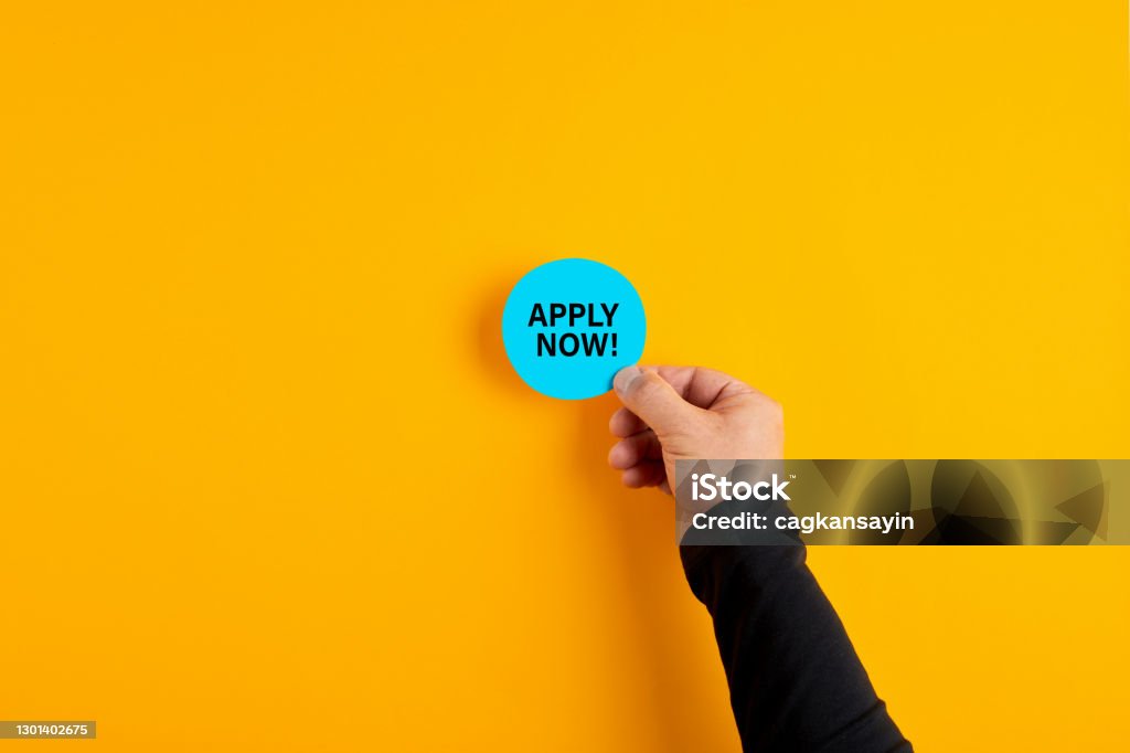Male hand holds a blue badge with the word apply now on yellow background. Male hand holds a blue badge with the word apply now on yellow background. Job vacancy, education or membership application concept. Applying Stock Photo
