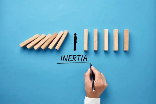 Businessman hand writing the word inertia with silhouette of a man standing against collapsing wooden dominos. Business inaction, apathy or inactivity concept.