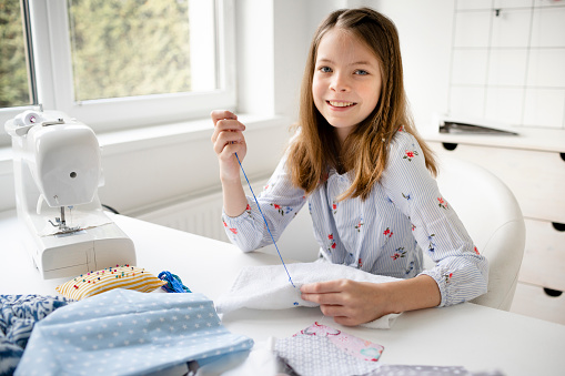 pretty and young schoolgirl is sitting by the table in her parents office embroidering, doing needlework