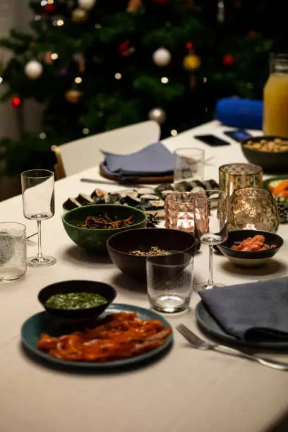 December 24, 2020 - Warsaw, Poland: 
Close up of a festive dinner table with Christmas tree in background. Linen tablecloths and napkins, candle holders, glasses, salmon dishes, sushi.