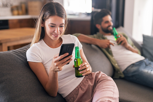 Very attractive women sitting at home with her boyfriend, smiling while holding mobile phone and beer