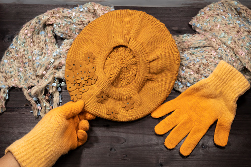On a dark background lies a beige scarf, a yellow knitted beret and a yellow glove. The hand in the second glove takes the beret.